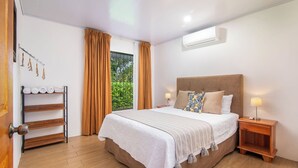 Een slaapkamer met een groot bed, een houten vloer, een raam met gordijnen, een nachtkastje met een lamp en een wandairconditioner.