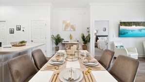 A dining area with a long table set for six, surrounded by chairs. There are potted plants on the walls and a television mounted on the wall.