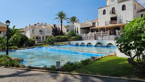 A swimming pool with a fountain, surrounded by greenery and a white building with a clock tower.