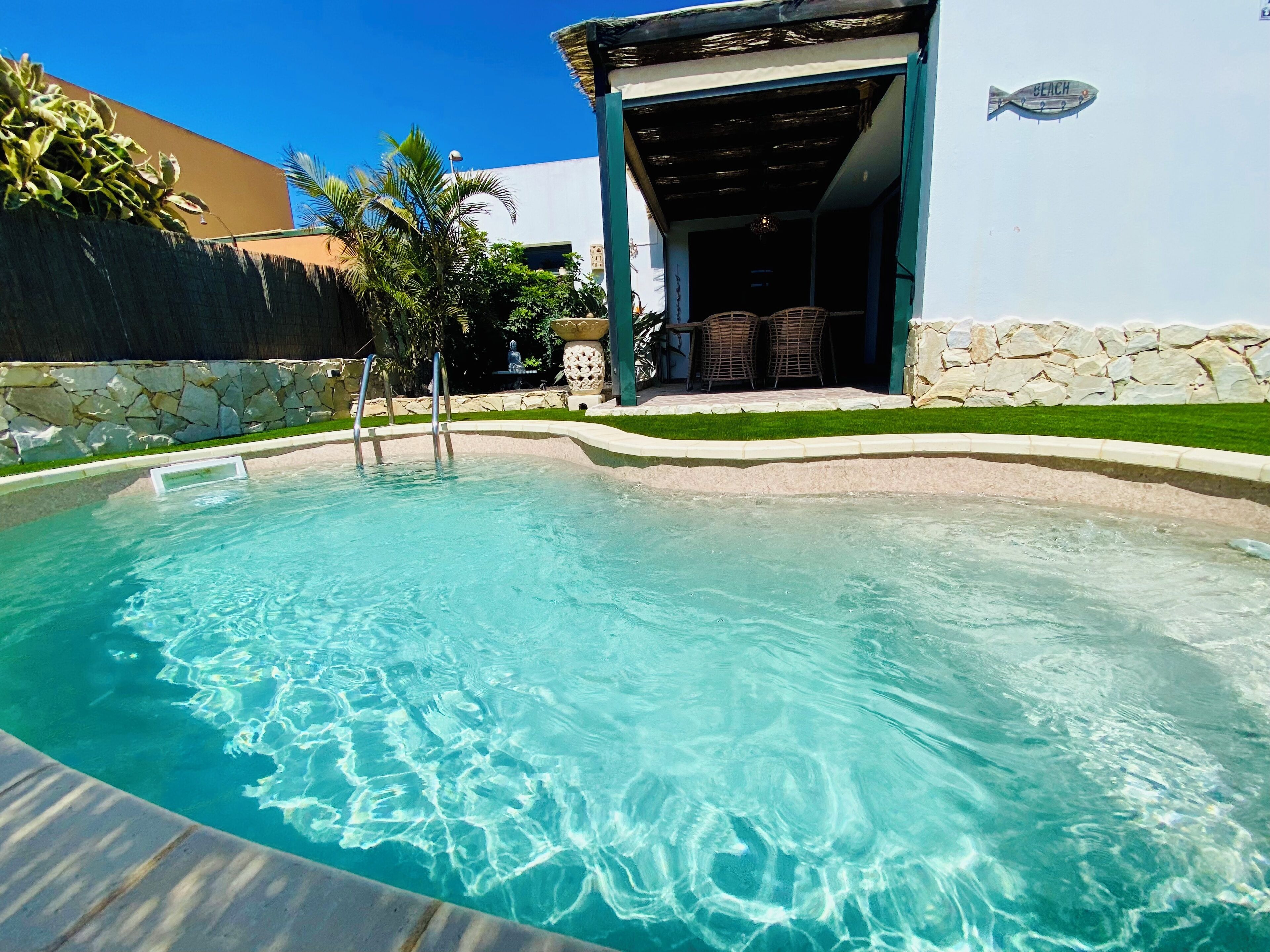 A clear swimming pool with a stone wall and a covered patio area.
