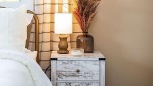 Un dormitorio con cama, mesita de noche con lámpara, florero con plantas secas y ventana con cortinas.