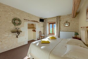 Quarto com uma cama grande, cabeceira de madeira, janela com cortinas, mesa pequena com abajur e televisão fixada na parede.