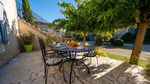 A stone patio with a round table and chairs, a potted plant, and a tree providing shade. - La Maison de Charlotte (Amberre)