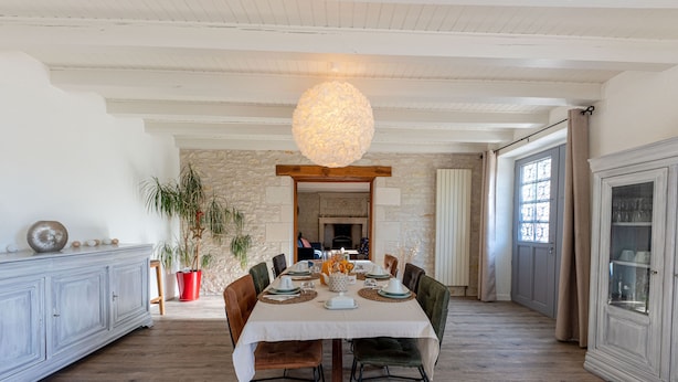 A dining area with a large table set for six, surrounded by wooden chairs with black metal frames. A unique spherical light fixture hangs from the ceiling, and a stone wall with a wooden archway is visible in the background. - La Maison de Charlotte (Amberre)