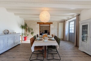 Uma sala de jantar com uma mesa grande posta para seis pessoas, rodeada por cadeiras de madeira com estrutura de metal preto. Um pendente esférico peculiar pendura-se do teto, e uma parede de pedra com um arco de madeira é visível ao fundo.