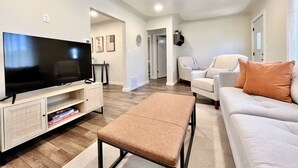 A modern living room with a flat-screen TV, a sofa, an ottoman, and a coffee table. There is a door leading to another room. - Cozy home. Newly remodeled and easy access to all that Grand Rapids has to offer (Grand Rapids)