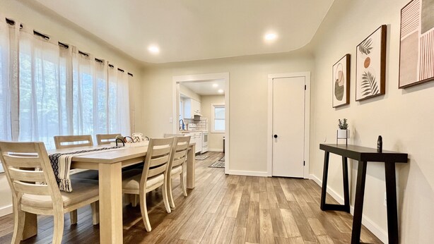 A dining area with a wooden table and chairs, a kitchen in the background, and a hallway with a door and framed pictures on the wall. - Cozy home. Newly remodeled and easy access to all that Grand Rapids has to offer (Grand Rapids)