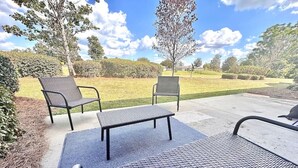 Un espace extérieur aménagé avec deux chaises et un banc placés sur un tapis texturé, le tout entouré d’un jardin paysager et d’arbres.