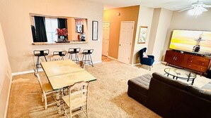 Un salon comprenant un canapé, une table basse, une table à manger avec des chaises, un tél éviseur et un coin cuisine avec des placards et une fenêtre.