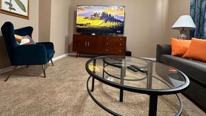 Un salon comprenant un canapé, un fauteuil bleu, une table basse en verre, un téléviseur posé sur un meuble en bois et une lampe.