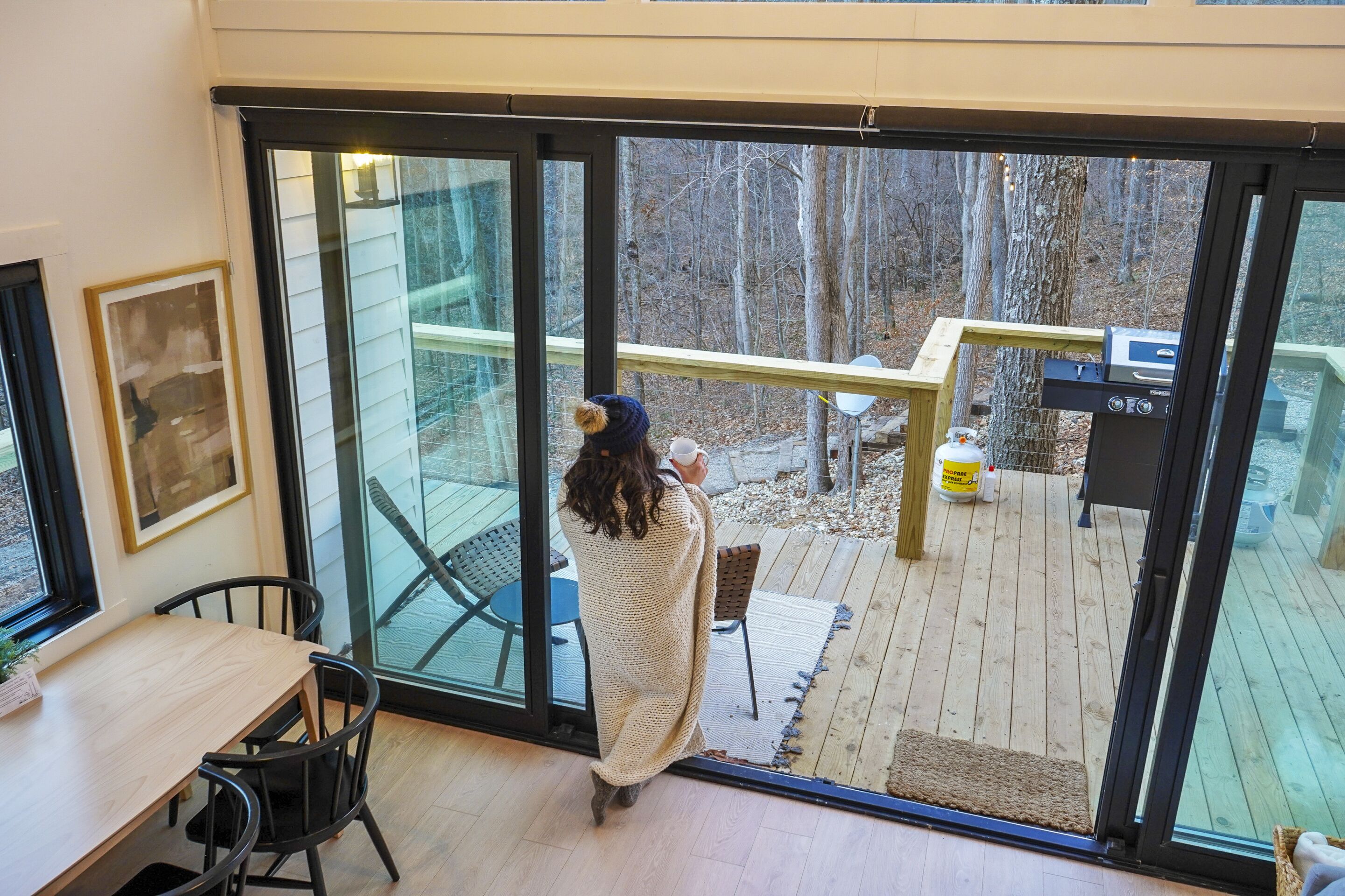 A person standing by a large glass door, looking outside. Inside, there is a wooden deck with outdoor furniture, a grill, and a water container.