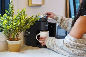 Una persona sosteniendo una taza blanca cerca de una máquina de café con una planta al lado.