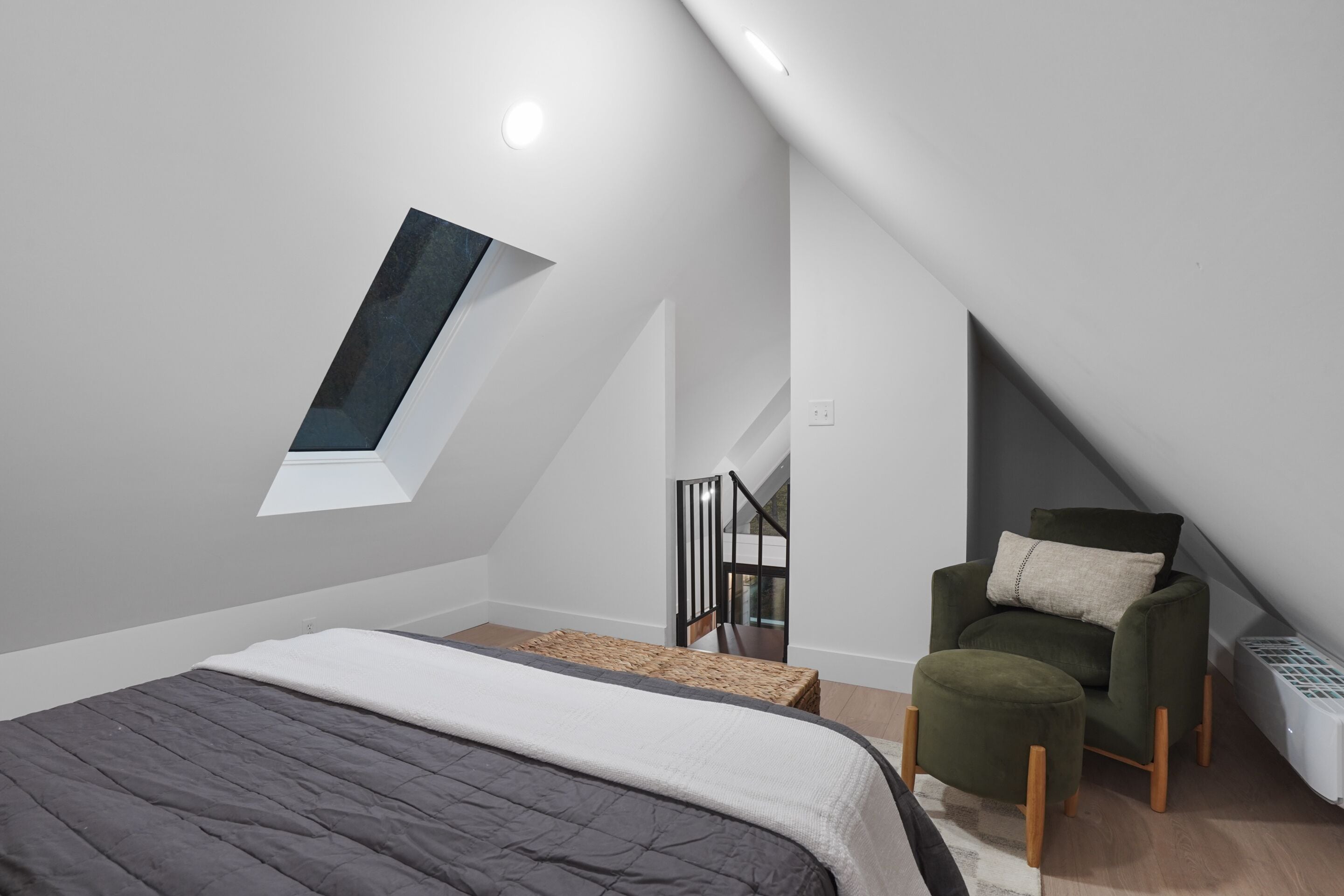 A modern loft bedroom with a sloped ceiling, a skylight, a bed with a grey quilt, a green armchair, and a staircase leading to an upper level.