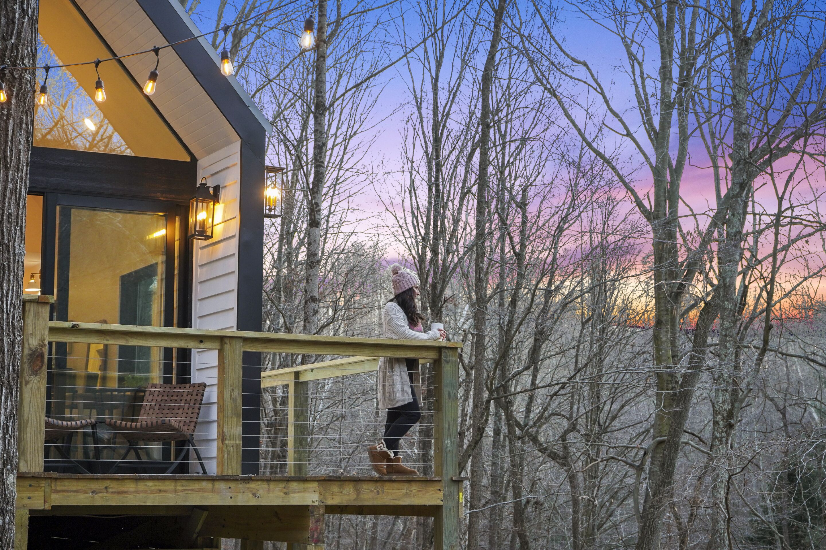 A wooden deck with a railing, a glass door leading to a room, and a view of trees against a sunset sky.