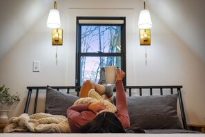 Une personne allongée sur un lit dans une pièce avec une fenêtre, deux lampes fixées au mur et une plante sur le côté.