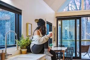 A woman sitting by a window with a cup, overlooking a dining area with chairs and a table.