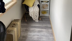 Un couloir étroit avec une fenêtre, une guirlande accrochée au mur, une étagère avec des livres et des objets décoratifs, une valise et une chaise recouverte d’une couverture.