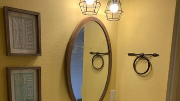 A bathroom with a yellow wall, a wooden vanity, a mirror with a wooden frame, two wall-mounted light fixtures, and two framed quotes.