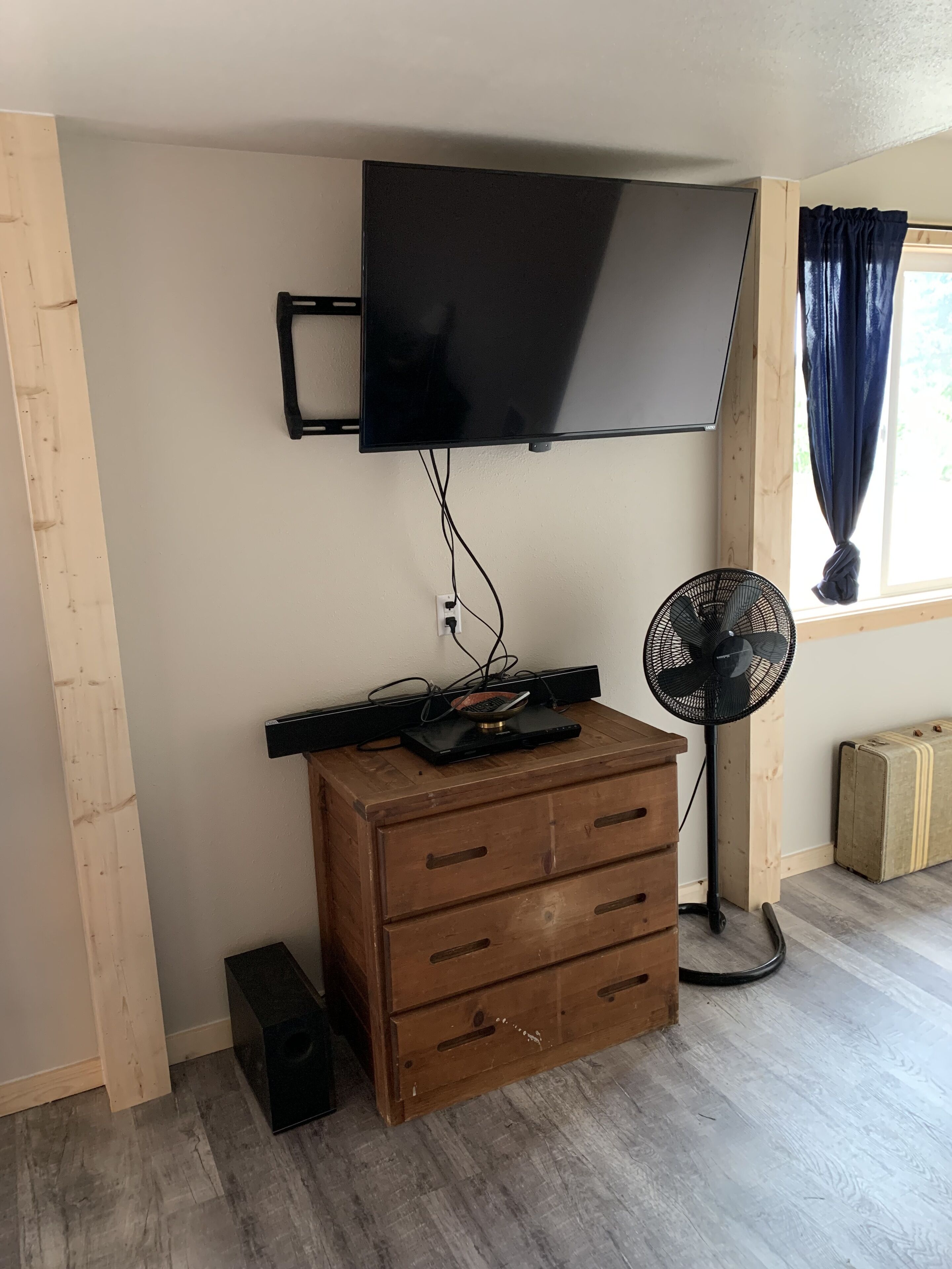 Een kamer met een wandtelevisie, een houten dressoir met lades, een ventilator en een raam met gordijnen.