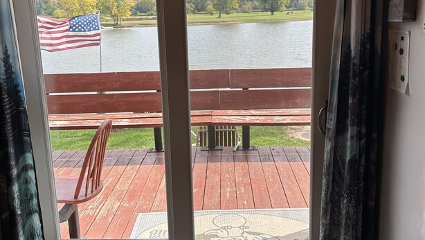 A room with a view of a lake, a wooden deck outside, and a rug with a logo.