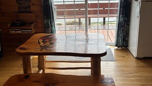 A wooden cabin interior with a wooden table and bench, a clock on the wall, and a view of a lake and mountains through a window.