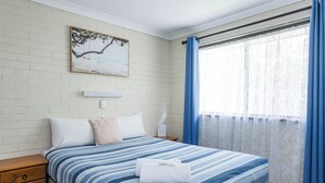Een slaapkamer met een bed, nachtkastje, houten dressoir, een aan de muur hangend schilderij en een raam met gordijnen.