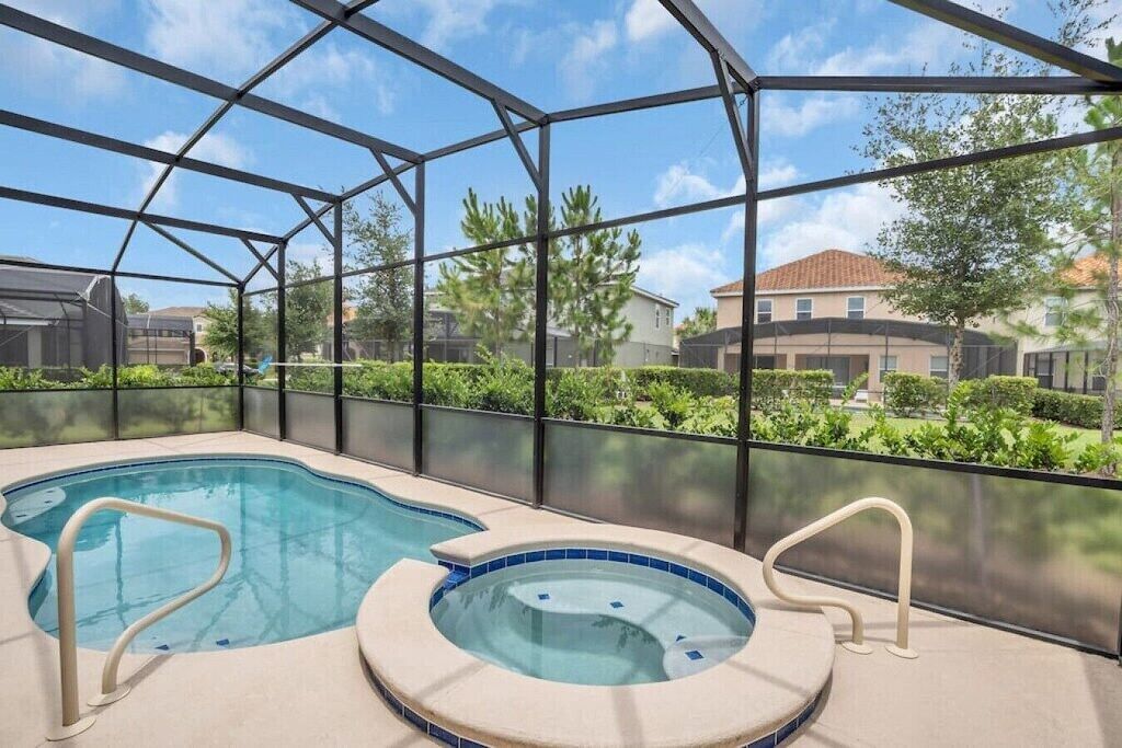 A swimming pool with a hot tub inside a covered area, surrounded by a metal frame and glass walls, with a view of houses and greenery outside.