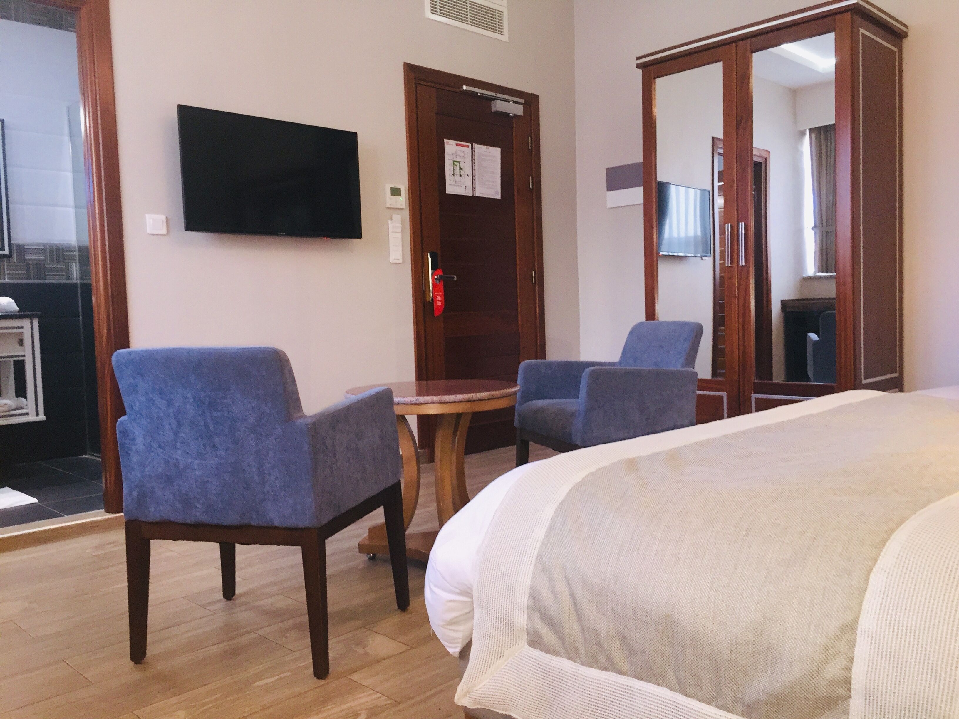 A hotel room with a bed, two armchairs, a small table, a flat-screen TV mounted on the wall, and a wooden wardrobe.