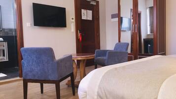 A hotel room with a bed, two armchairs, a small table, a flat-screen TV mounted on the wall, and a wooden wardrobe.