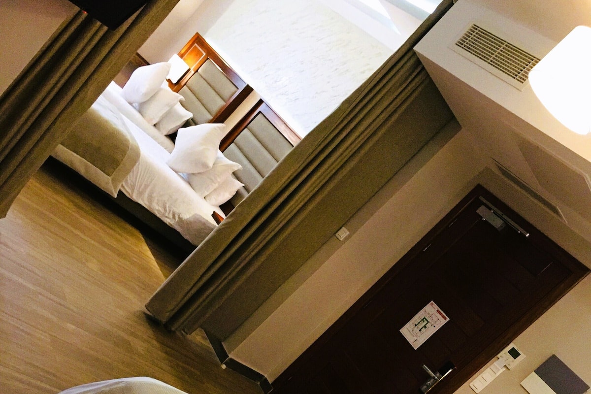 A hotel room with two beds, a wooden floor, a dark door, and a window with curtains.