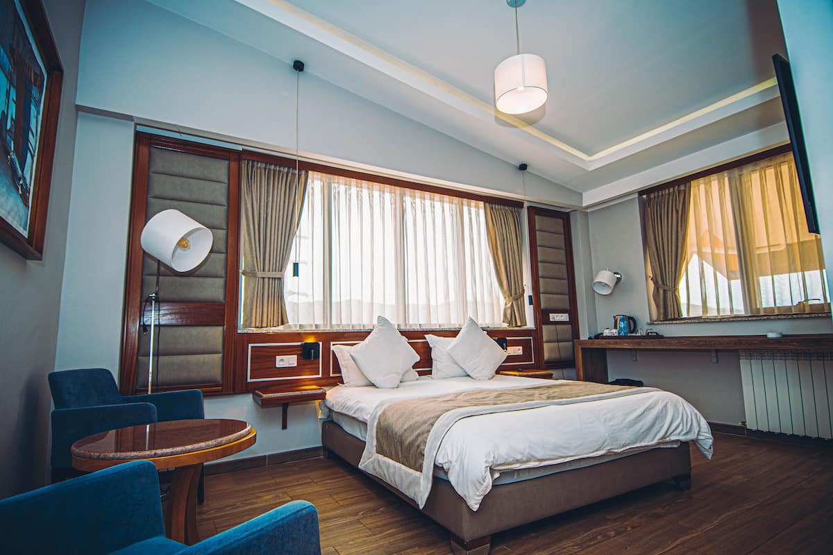 A hotel room with a large bed, two bedside tables, a round wooden table, two blue armchairs, and a large window with curtains.