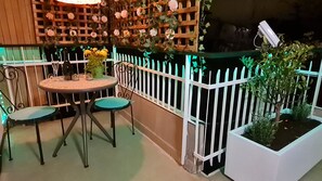 Een terras met een ronde tafel en twee stoelen, een witte picket fence en potplanten.