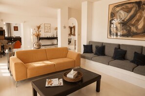 A living room with a sofa and a coffee table, a dining area with chairs and a table, and a kitchen in the background. - Mas des Bécasses in Provence 22 pax (Arles)