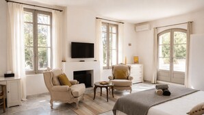 A bedroom with a bed, two armchairs, a small table, a fireplace, a TV, and large windows. - Mas des Bécasses in Provence 22 pax (Arles)