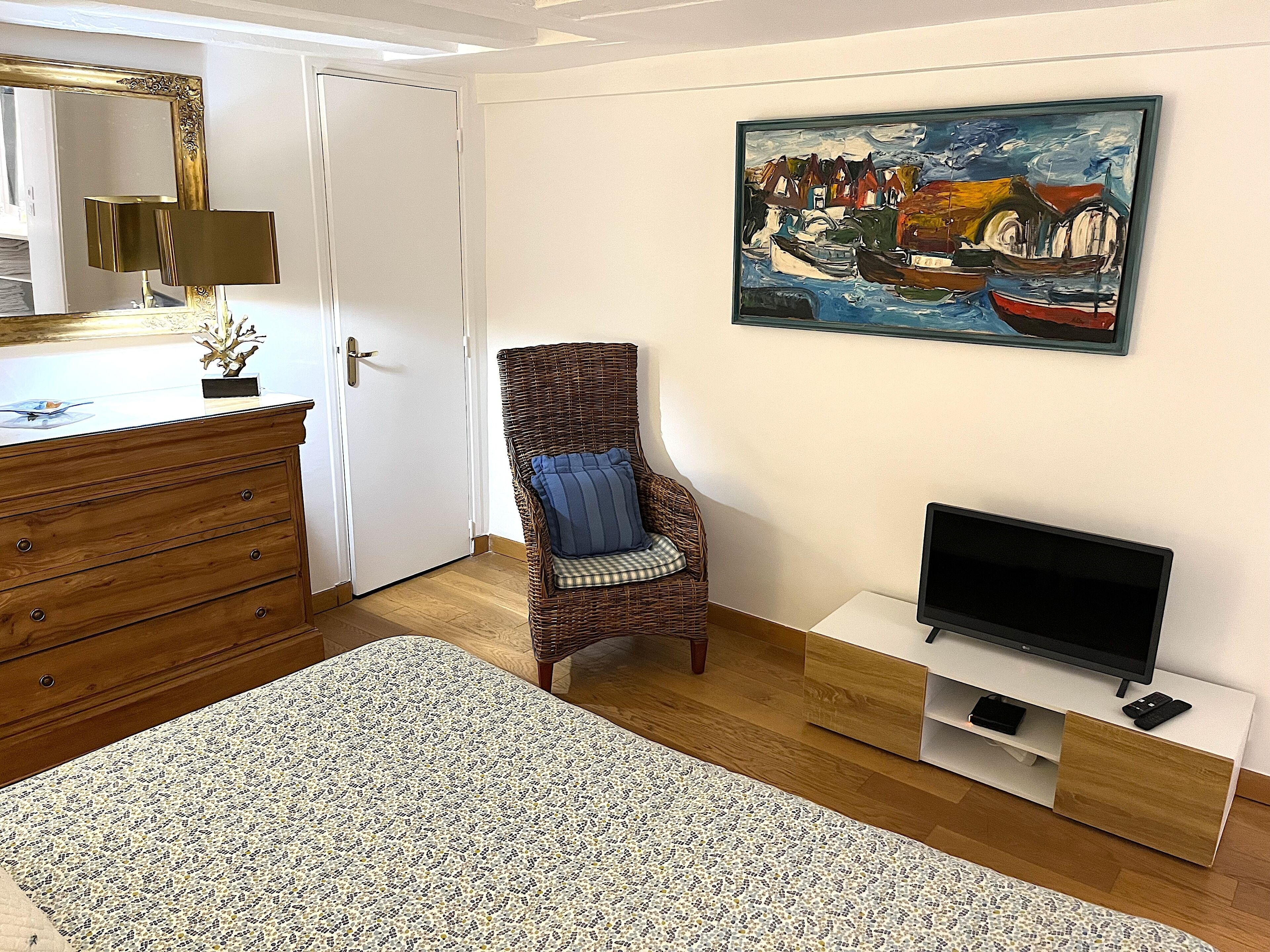 A room with a wooden dresser, a wicker chair with a blue cushion, a television on a white stand, and a painting of a harbor scene on the wall.