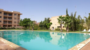 A swimming pool with a clear blue water surface, surrounded by palm trees and a building with multiple balconies. - Dar Lala Mama (Marrakech)