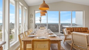 A dining area with a wooden table set for six, surrounded by wicker chairs, a sofa, and a large window offering a view of the ocean. - Brand New Luxury Beach Home w Hot Tub & Sauna (Rockaway Beach)