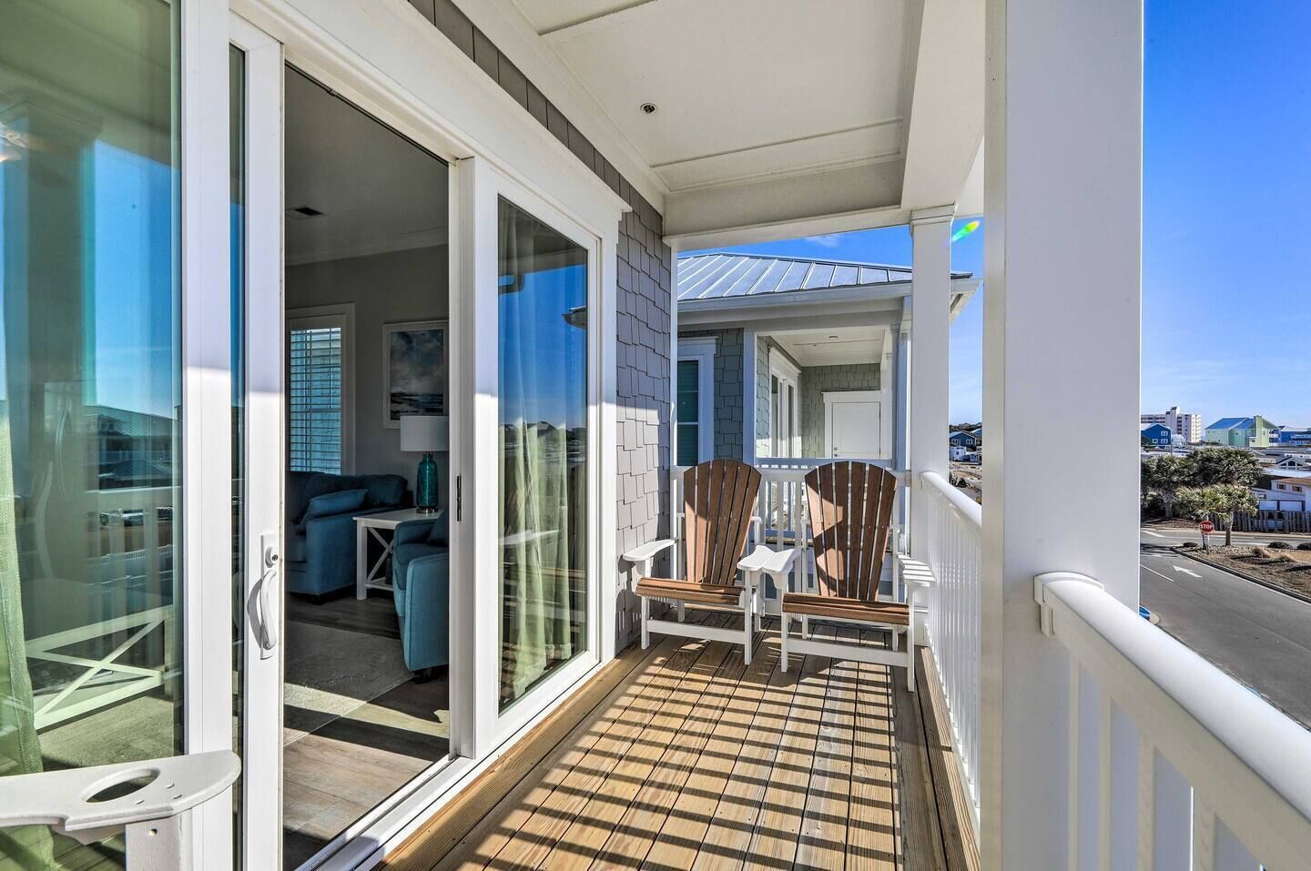 A balcony with a wooden deck, sliding glass doors leading to an interior with a sofa and a table, and a view of the street and buildings in the background.