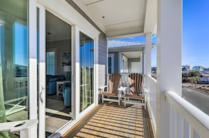 Een balkon met een houten terras, schuiframen die uitkomen op een interieur met een bank en een tafel, en een uitzicht op de straat en gebouwen op de achtergrond.