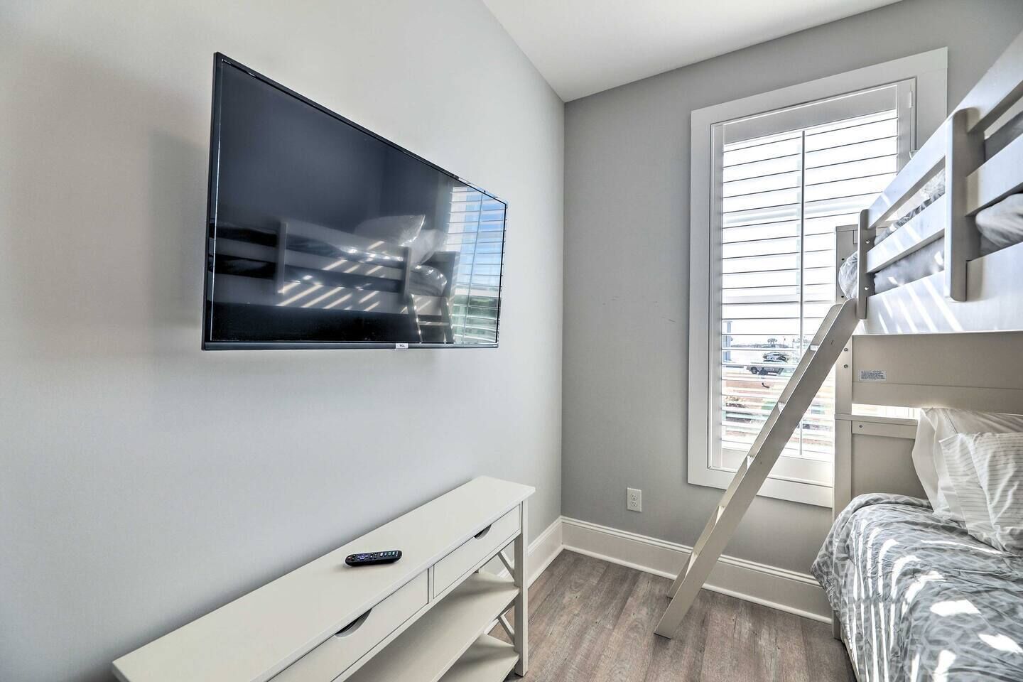 A bedroom with a TV mounted on the wall, a bunk bed, a desk with a chair, and a window with blinds.