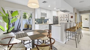 A modern kitchen with white cabinets, a central island, and bar stools. There are potted plants and a dining table with chairs.