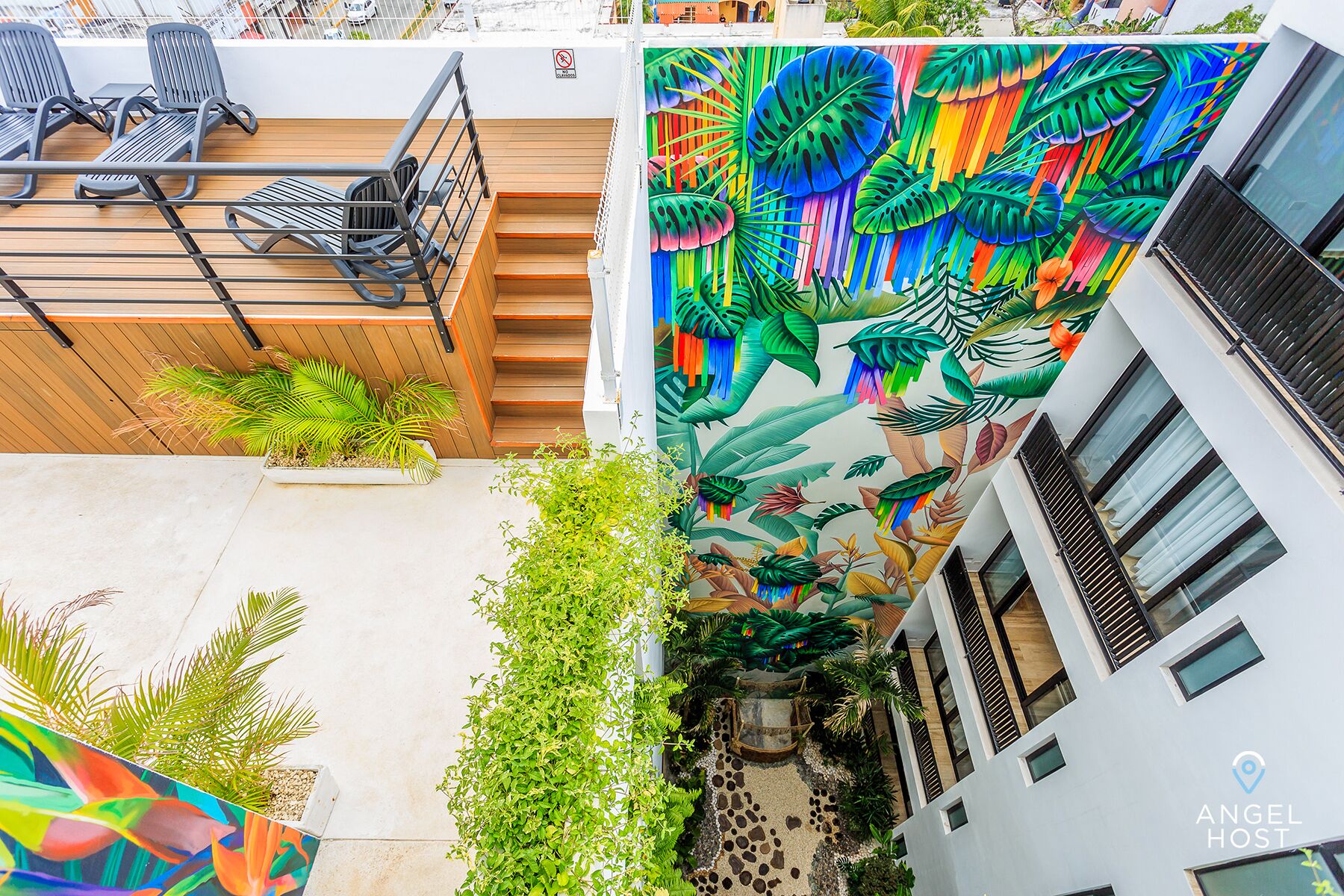 Un edificio moderno con un mural vibrante de hojas y flores tropicales, mobiliario exterior con sillas y mesas, y un camino de piedra.
