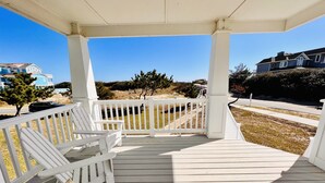A covered porch with white railings and a bench, overlooking a beach with sparse vegetation and houses in the background. - *NEW -25ft to Beach, 2br. Oceanviews.Pool.Hot Tub (Corolla)
