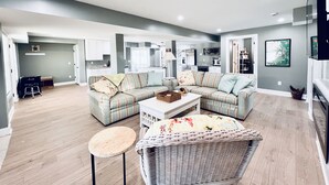 A spacious living room with a large sectional sofa, a small round table, and a wicker chair. - *NEW -25ft to Beach, 2br. Oceanviews.Pool.Hot Tub (Corolla)