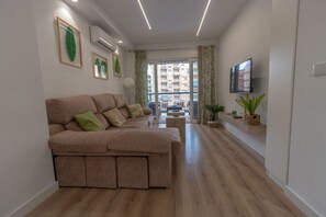 Een moderne woonkamer met een hoekbank, een flatscreen televisie en een groot raam met uitzicht op een gebouw.