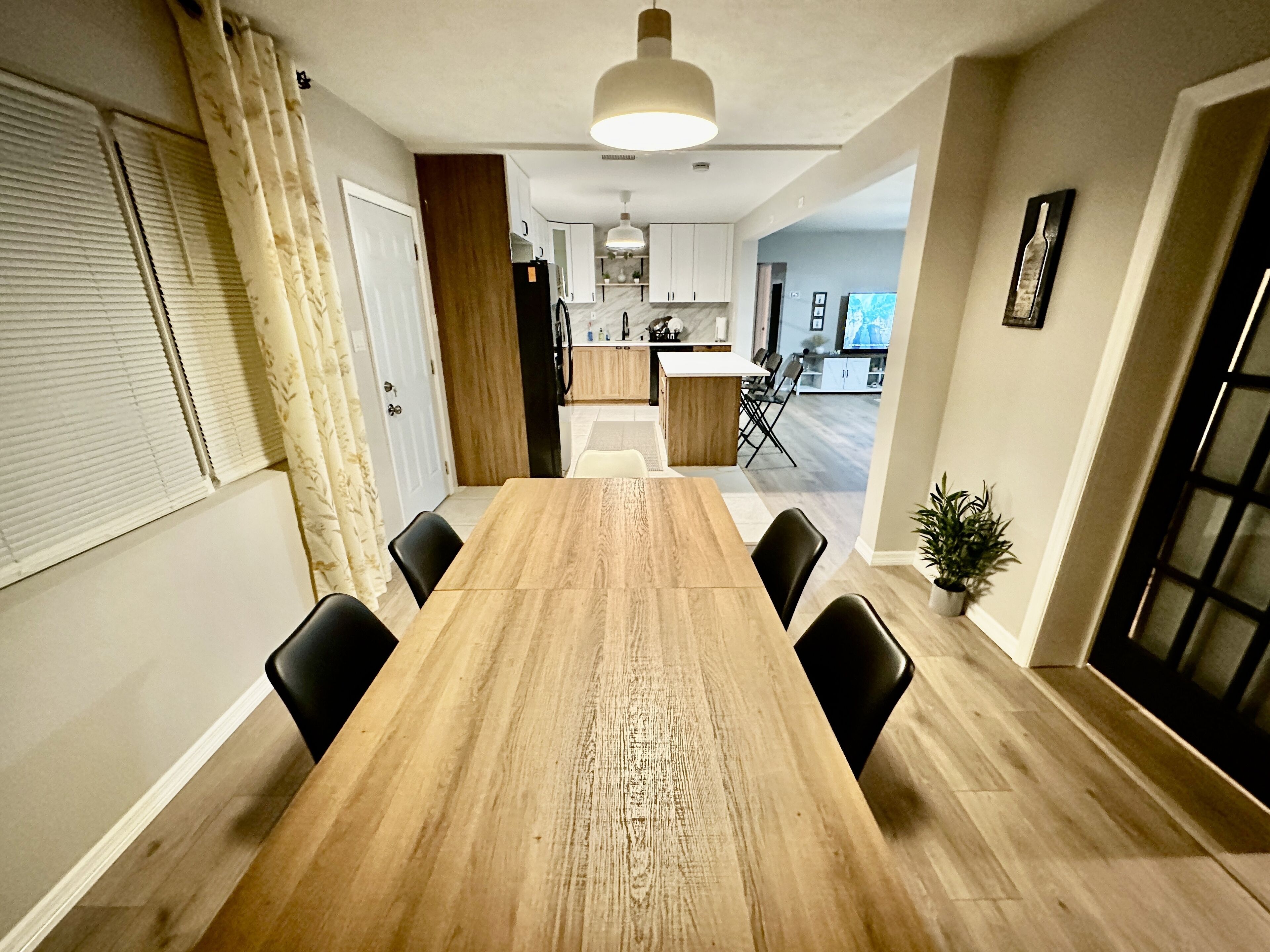 Un coin repas avec une table en bois et des chaises noires, une cuisine en arrière-plan et une plante en pot posée sur le sol.