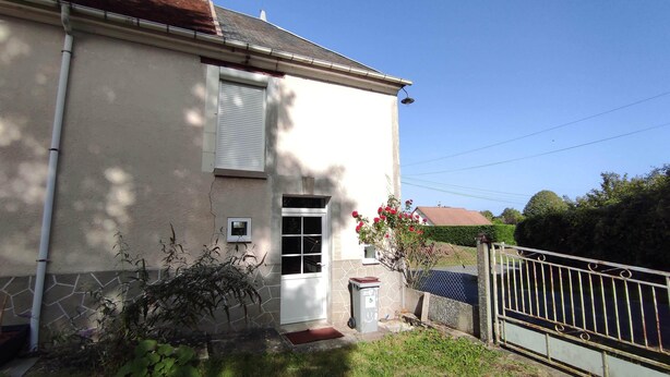 A two-story house with a white exterior, a front door, and a small window. There is a metal gate and a red flower bush in the front yard. - Gîte Indépendant à la Campagne (Couffy)