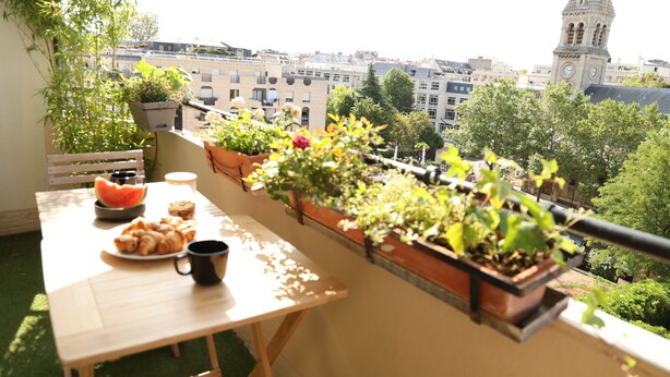 Ein Balkon mit Tisch, Blumentöpfen und Blick auf eine Stadtlandschaft, darunter ein Kirchturm.