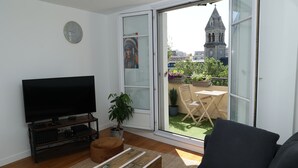 Ein Wohnzimmer mit Flachbildfernseher, einer Couch, einem Holztisch und einer Glastür, die auf einen Balkon mit Blick auf einen Kirchturm führt.
