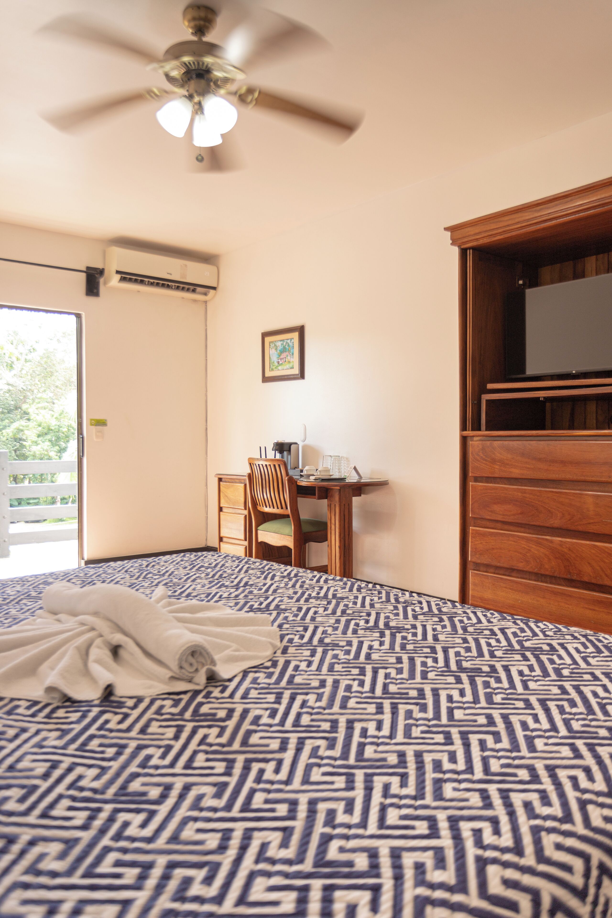 A bedroom with a patterned bedspread, a ceiling fan, a desk with a chair, a television, and a wardrobe.